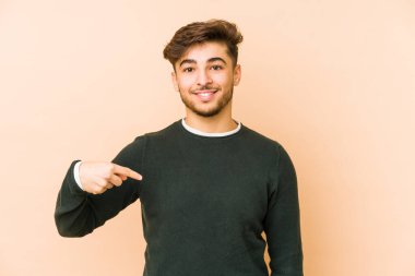 Bej bir arka planda izole edilmiş genç bir Arap adam gömlek fotokopi alanını işaret ediyor, gururlu ve kendinden emin.