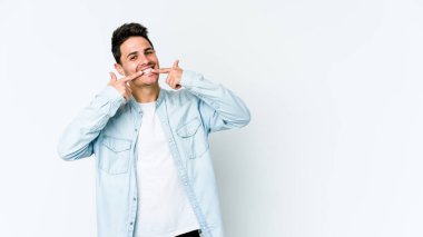 Beyaz arka planda izole edilmiş genç beyaz adam gülümsüyor, parmaklarını ağıza doğrultuyor..