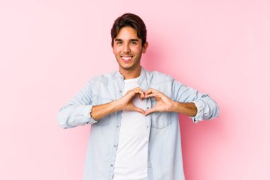 Pembe arka planda poz veren beyaz bir genç izole edilmiş gülümseyen ve elleriyle kalp şeklini gösteren bir adam..