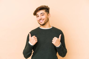 Genç Arap bir adam bej bir arka planda izole edilmiş. Baş parmağını kaldırıyor, gülümsüyor ve kendinden emin..