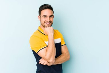 Mavi arka planda izole edilmiş genç beyaz adam gülümsüyor ve kendinden emin, elleriyle çenesine dokunuyor..