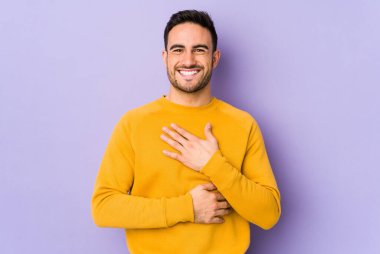 Mor arka planda izole edilmiş genç beyaz adam mutlu bir şekilde gülüyor ve ellerini karnının üstünde tutarak eğleniyor..