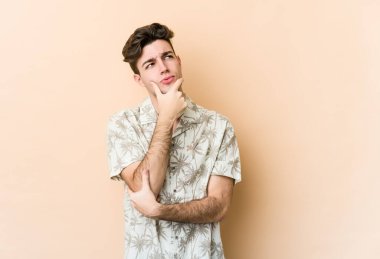 Beyaz tenli genç adam bej arka planda izole edilmiş düşünüyor ve yukarı bakıyor, yansıtıyor, düşünüyor, bir fantezi kuruyor..