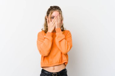 Beyaz arka plandaki genç beyaz kadın kameraya parmaklarıyla göz kırpıyor, utanarak yüzünü kapatıyor..