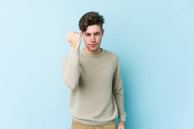 Mavi arka planda izole edilmiş genç beyaz adam yumruğunu kameraya gösteriyor, agresif yüz ifadesi var..