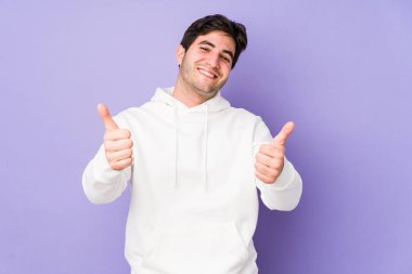 Mor arka planda izole edilmiş genç adam gülümsüyor ve baş parmağını kaldırıyor.