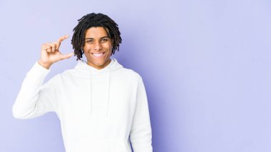 Genç Afrikalı Amerikalı Rasta adam işaret parmaklarıyla küçük bir şey tutuyor, gülümsüyor ve kendinden emin..