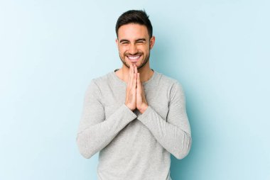 Mavi arka planda izole edilmiş genç beyaz bir adam el ele tutuşup, ağzının kenarında dua ediyor, kendinden emin..