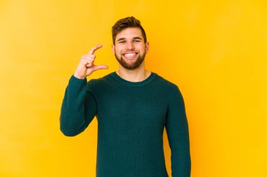 Sarı arka planda izole edilmiş genç beyaz adam işaret parmaklarıyla küçük bir şey tutuyor, gülümsüyor ve kendinden emin..