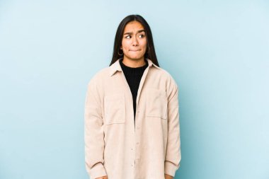 Mavi bir arka planda izole edilmiş genç bir kadın kafası karışmış, şüpheli ve güvensiz hissediyor..