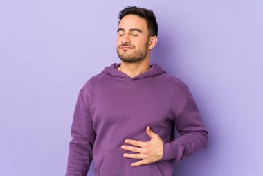 Mor arka planda izole edilmiş genç beyaz adam karnına dokunuyor, nazikçe gülümsüyor, yemek yiyor ve tatmin oluyor..