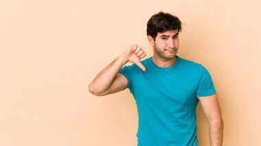 Bej arka planda izole edilmiş genç adam baş parmağını aşağı gösteriyor, hayal kırıklığı konsepti..