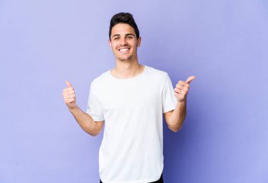 Mor arka planda izole edilmiş genç beyaz adam iki parmağını da kaldırıyor, gülümsüyor ve kendinden emin..