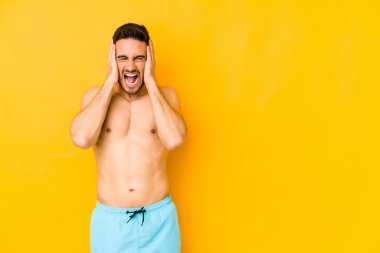 Sarı arka planda mayosu olan genç, beyaz bir adam. Kulakları çınlayan, elleri kulakları çınlayan, yüksek ses duymamaya çalışan beyaz bir adam..