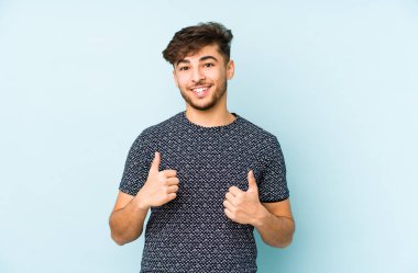 Genç Arap bir adam mavi bir arka planda izole edilmiş. Baş parmağını kaldırıyor, gülümsüyor ve kendinden emin..