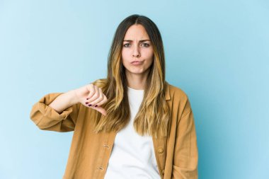 Mavi arka planda izole edilmiş genç beyaz kadın baş parmağını aşağı gösteriyor, hayal kırıklığı konsepti.