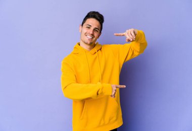 Mor arka planda izole edilmiş genç beyaz bir adam. Elinde işaret parmaklarıyla küçük bir şey tutuyor, gülümsüyor ve kendinden emin..