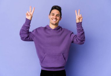 Mor arka planda izole edilmiş genç beyaz adam zafer işareti gösteriyor ve gülümsüyor..