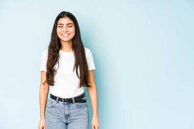 Mavi arka plandaki genç Hintli kadın gülüyor ve gözlerini kapatıyor, rahatlamış ve mutlu hissediyor..