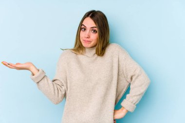 Kafkasyalı genç bir kadın mavi arka planda izole edilmiş sorgulama sırasında omuz silkiyor ve kuşkulanıyor..