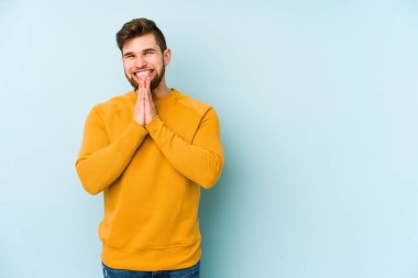 Mavi arka planda izole edilmiş genç beyaz bir adam el ele tutuşup, ağzının kenarında dua ediyor, kendinden emin..