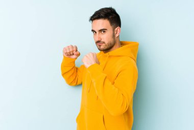 Kafkasyalı genç bir adam mavi arka planda izole edilmiş. Yumruk atıyor, sinirleniyor, kavga ediyor, kavga ediyor..