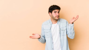 Bej arka planda izole edilmiş genç bir adam. Kafası karışmış ve omuz silkiyor. Kopya alanını tutmak için..