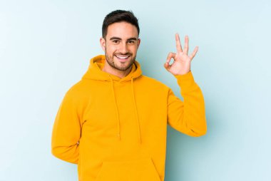 Kafkasyalı genç bir adam mavi arka planda izole edilmiş bir şekilde göz açıp kapayana kadar iyi bir el hareketi yapıyor..