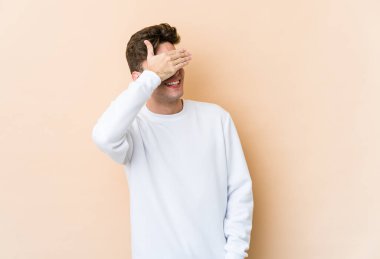 Beyaz tenli, bej arka planda izole edilmiş genç bir adam elleriyle gözlerini kapatıyor, gülümseyerek büyük bir sürpriz bekliyor..