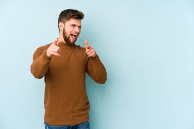 Mavi arka planda izole edilmiş genç beyaz adam parmaklarıyla ön tarafı işaret ediyor..