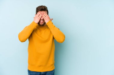 Mavi arka planda soyutlanmış genç beyaz bir adam elleriyle gözlerini kapatıyor, geniş bir gülümsemeyle bir sürpriz bekliyor..