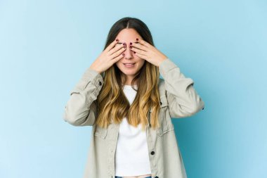 Mavi arka planda izole edilmiş genç beyaz kadın elleriyle gözlerini kapatmaktan korkuyor..