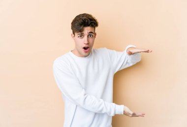 Beyazlı genç bir adam bej arka planda izole edilmiş. Elleri arasında bir kopya alanı tutarken şok olmuş ve hayrete düşmüş..