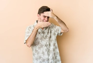 Beyazlı genç bir adam bej arka planda izole edilmiş. Kameraya parmaklarıyla göz kırpıyor, yüzünü kapatmaktan utanıyor..