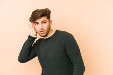 Genç Arap bir adam bej bir arka planda izole edilmiş bir şekilde bir dedikoduyu dinlemeye çalışıyor..