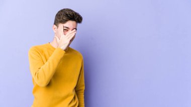 Mor arka planda izole edilmiş genç beyaz adam kameraya parmaklarıyla göz kırpıyor, yüzünü kapatmaktan utanıyor..