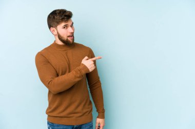Mavi arka planda izole edilmiş genç beyaz adam gülümsüyor ve boş alanda bir şeyler gösteriyor..