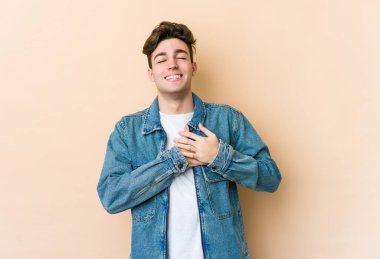 Beyazlı genç bir adam bej arka planda izole edilmiş. Gülüyor, elini kalbe koyuyor, mutluluk kavramını koruyor..