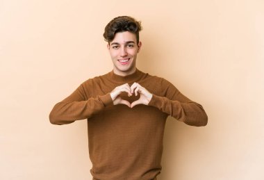 Beyaz tenli genç adam bej arka planda izole edilmiş gülümsüyor ve elleriyle kalp şeklini gösteriyor..