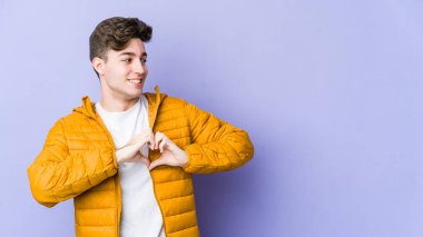 Mor arka planda izole edilmiş genç beyaz adam gülümsüyor ve elleriyle kalp şeklini gösteriyor..