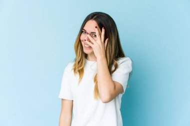 Mavi arka planda izole edilmiş genç beyaz kadın kameraya parmaklarıyla göz kırpıyor, yüzünü kapatmaktan utanıyor..