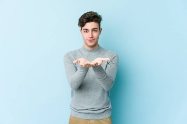 Kafkasyalı genç adam mavi arka planda izole edilmiş. Elinde avuç içi olan bir şey var ve kameraya sunuyor..