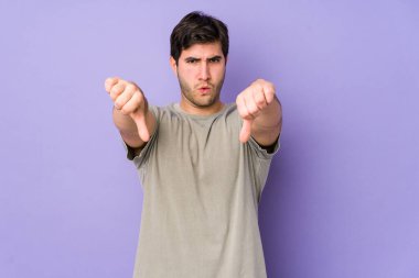 Mor arka planda izole edilmiş genç bir adam baş parmağını aşağı gösteriyor ve hoşlanmadığını ifade ediyor..