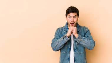 Genç adam bej arka planda izole edilmiş şans için dua ediyor, şaşırmış ve ağzı açık bir şekilde önüne bakıyor..