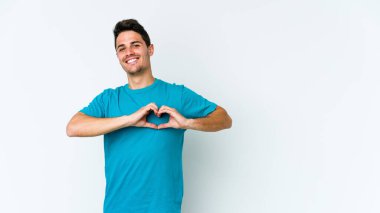 Beyaz arka planda izole edilmiş genç beyaz bir adam gülümsüyor ve elleriyle kalp şekli gösteriyor..