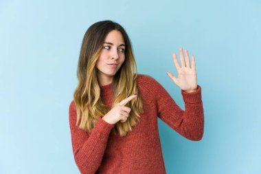 Mavi arka planda izole edilmiş genç beyaz kadın gülümseyerek beş numarayı parmaklarıyla gösteriyor..