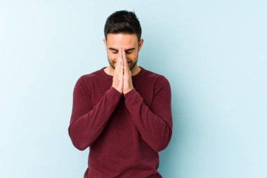 Mavi arka planda izole edilmiş genç beyaz bir adam el ele tutuşup, ağzının kenarında dua ediyor, kendinden emin..