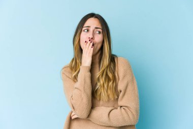 Mavi arka planda izole edilmiş genç beyaz kadın esniyor. Ağzı eliyle kapatan yorgun bir hareket sergiliyor..