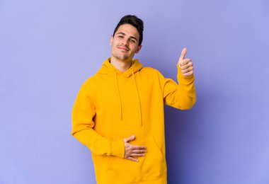 Mor arka planda izole edilmiş genç beyaz adam karnına dokunuyor, nazikçe gülümsüyor, yemek yiyor ve tatmin oluyor..