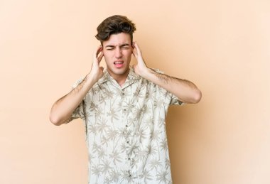 Beyazlı genç bir adam bej arka planda izole edilmiş parmaklarıyla kulaklarını kapatıyor, gürültülü bir ortam yüzünden stresli ve çaresiz..